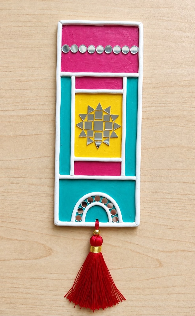 Pustaka — Handcrafted Lippan Keepsake Bookmark with Silk Tassel — handmade hand-piped clay relief, celestial mirror starburst, and celebratory color blocks on wooden plaque, with red silk tassel by Rachna Saxena